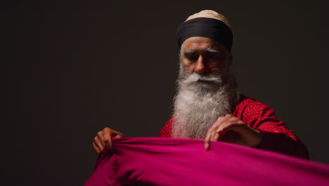 Fotografía-De-Estudio-Con-Iluminación-Tenue-De-Un-Hombre-Sikh-Mayor-Doblando-Tela-Para-Un-Turbante-Contra-Un-Fondo-Oscuro-Liso-3